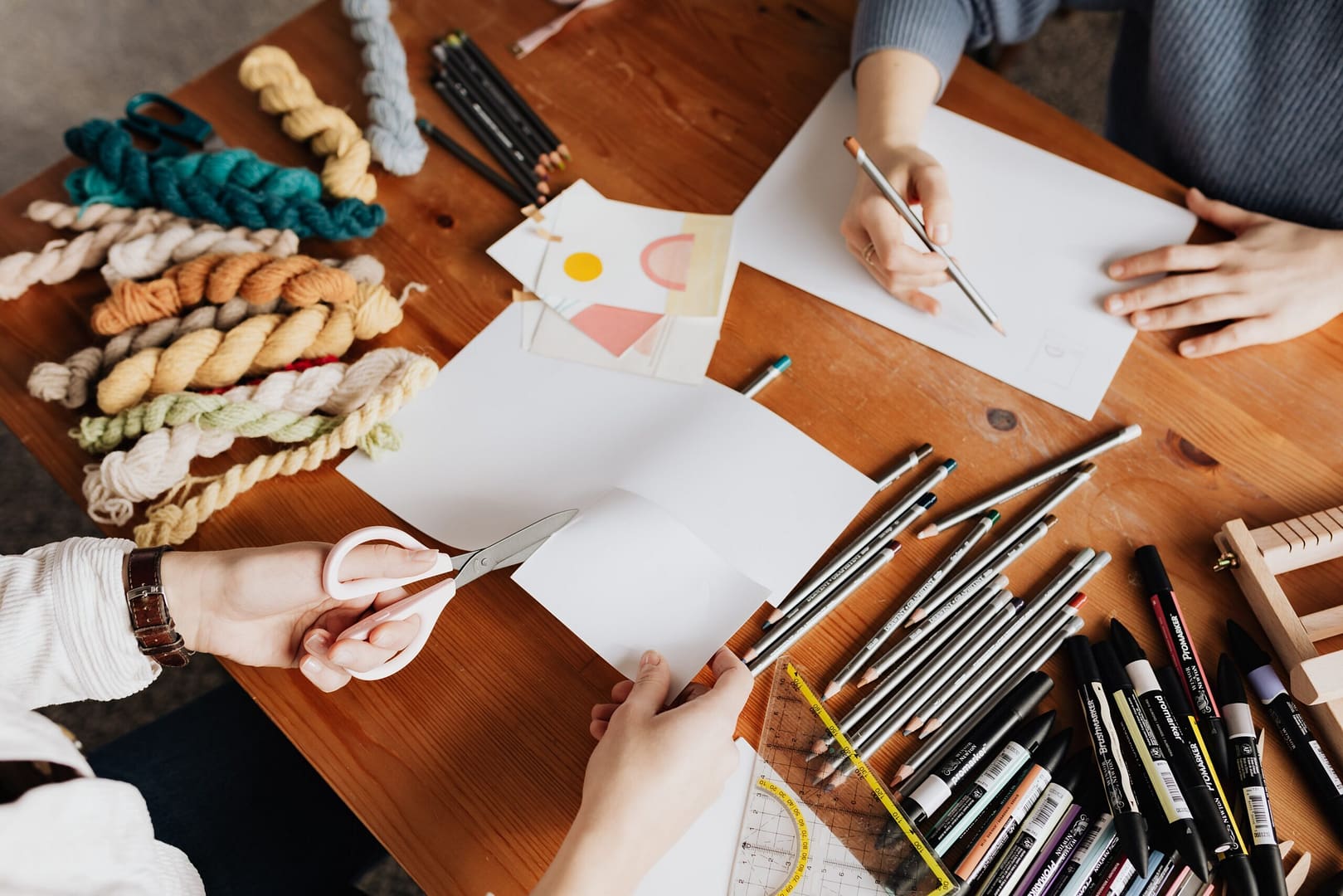 students doing arts and crafts , one drawing and one cutting paper with pencils and cloth scattered around